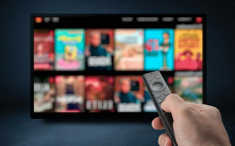 Hand holding a remote control in front of a smart TV streaming movies, TV shows, and on-demand entertainment content.